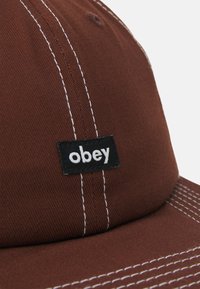 Hnedá čiapka vyrobená z tkaniny s bielymi švy. Obsahuje čiernu obdĺžnikovú nálepku s nápisom "obey" v bielej farbe.