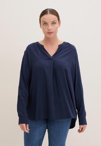 Blusa de manga larga azul marino con escote en V, corte holgado, textura suave y dobladillo curvado, combinada con jeans azules.