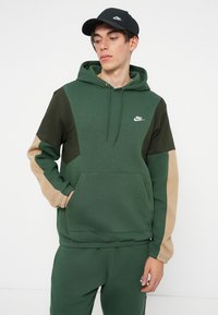 Vihreä huppari, jossa on tummanvihreät ja vaaleanruskeat väriblokatut hihat. Siinä on kengurutasku ja nyöritettävä huppu. Nike-logo rinnassa.