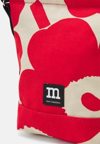 Rød lærredstaske med stort blomstermotiv i rød og creme, med en sort logo-patch. Holdbart materiale og struktureret form.