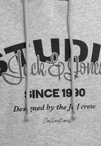 Šedá mikina s kapucí a stahovacími šňůrkami, na které je výrazný černý nápis "STUDI" a "Jack & Jones", s detailním písmem a ležérním designem.