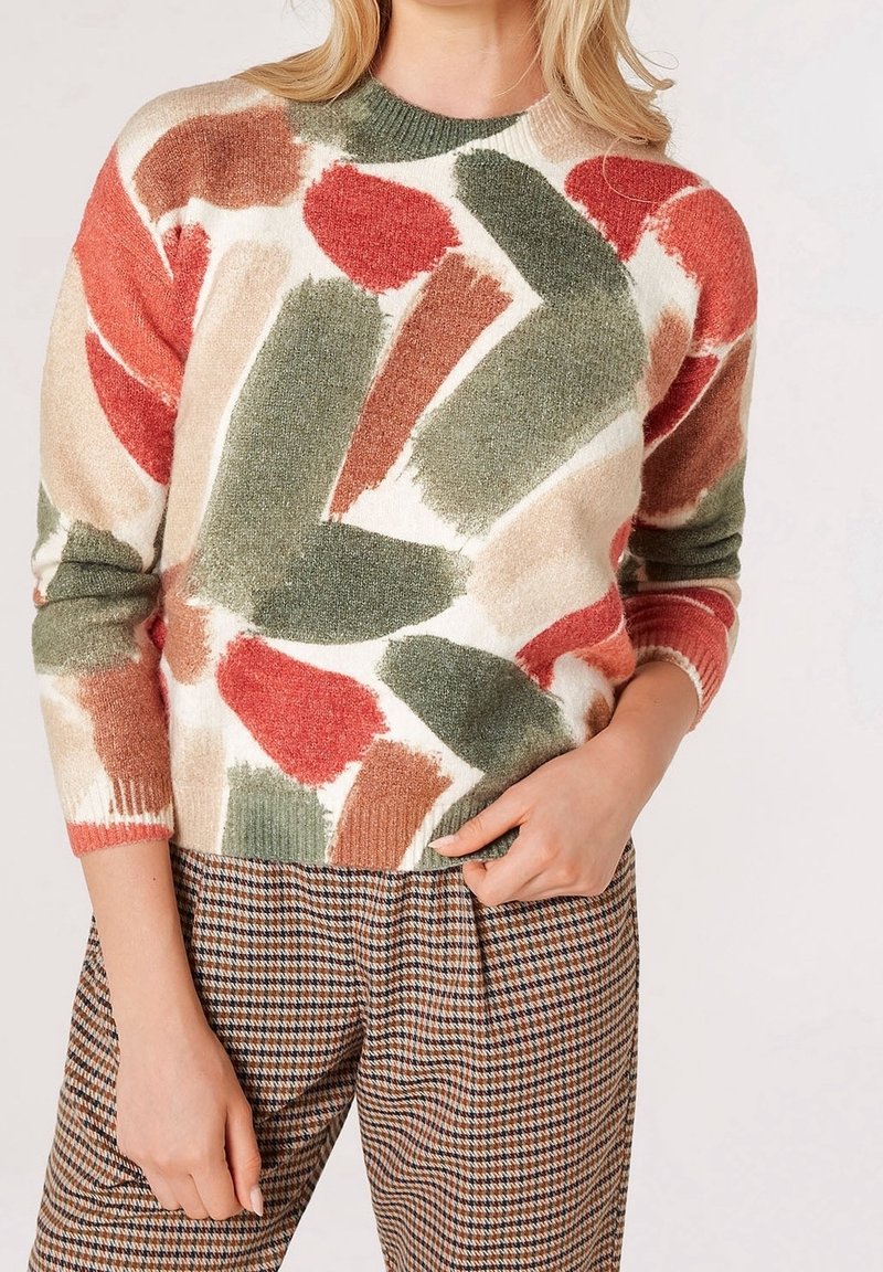 Knit sweater featuring a multicolored abstract pattern in green, red, and beige. Ribbed neckline and cuffs. Paired with checked trousers.