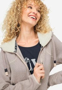 Chaqueta con capucha de cierre éclair en gris claro, con un collar de piel sintética, cordones de contraste y una textura suave; llevada sobre una camiseta gráfica azul marino.