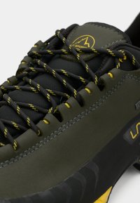 Μαύρα και ελαιόλαδα μαθήματα αναρρίχησης με υφές, κίτρινες λεπτομέρειες και μοτίβο στα κορδόνια. Διαθέτει ενισχυμένα μάτια και ξεχωριστή υφασμάτινη σόλα.