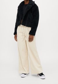 Schwarze Kunstfell-Cropped-Jacke mit breitem Kragen, kombiniert mit cremefarbenen Corduroy-weiten Hosen und schwarzen Sneakers mit weißen Sohlen.