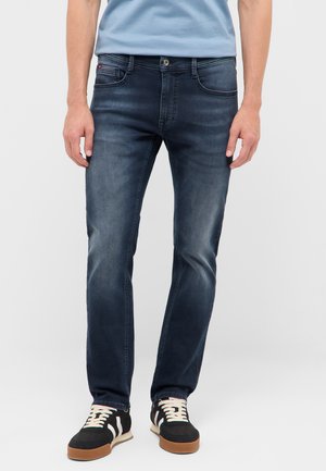 Person trägt dunkelblaue Slim-Fit-Jeans, hellblaues T-Shirt und schwarze Sneaker mit weißen Schnürsenkeln und steht vor einem einfarbigen Hintergrund.