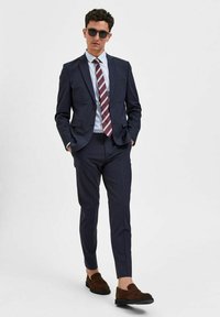 Navy pinstripe dressjakke med skreddersydd passform, lyseblå stripete skjorte, maroon og hvit stripete slips, og brune loafers. Solbriller inkludert.