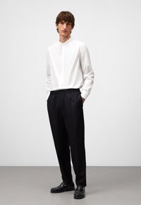 Camicia bianca a maniche lunghe con colletto alla cinese, abbinata a pantaloni neri larghi e scarpe in pelle nere, su uno sfondo semplice.