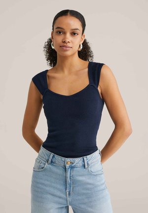 Femme aux cheveux bouclés portant des boucles d'oreilles argentées en anneau, un haut sans manches côtelé bleu marine et un jean taille haute bleu clair, debout les mains dans le dos.