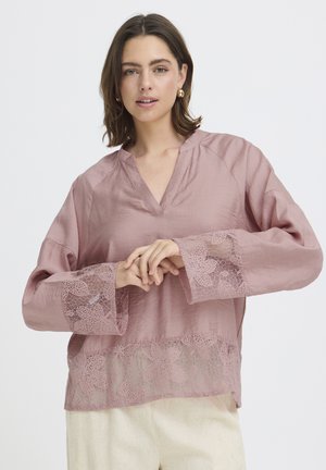 Femme aux cheveux bruns portant un chemisier rose clair avec des détails en dentelle florale aux poignets et à l'ourlet, associé à un pantalon crème, debout sur un fond blanc.