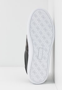 Semelle d'une basket blanche avec des logos à motifs et une étiquette rectangulaire "GUESS" au centre, placée à la verticale contre un mur.