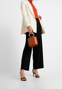Blazer crème porté sur un haut orange, associé à un pantalon large noir. Un sac à main marron texturé avec une anse ronde complète la tenue.