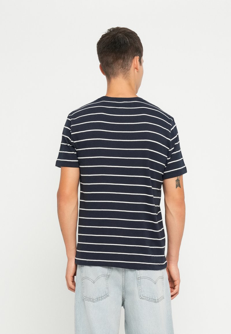 Niebieska koszulka w paski z białymi poziomymi paskami, krótkie rękawy, okrągły dekolt i miękka tekstura, noszona z jasnymi dżinsowymi szortami.