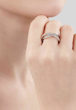 Primo piano di una mano con un anello d'argento intrecciato sul dito medio che tocca il mento su uno sfondo neutro.