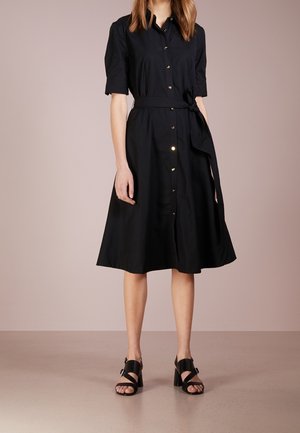 Robe noire à manches courtes, avec ceinture à la taille et jupe évasée ; présente des boutons dorés et une texture en coton lisse. Des chaussures noires complètent la tenue.