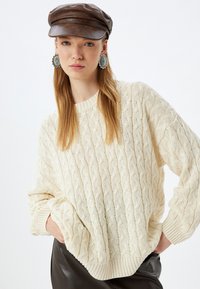 Cremfarbener, grob gestrickter Pullover mit lockerer Passform, Rippbündchen und Saum, kombiniert mit einer braunen Lederkappe und großen runden Ohrringen.