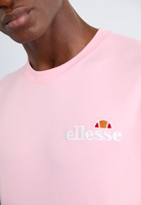 Svijetloružičasta pamučna majica s okruglim izrezom i bijelim izvezenim 'ellesse' logom s šarenim motivom iznad. Glatka tekstura.