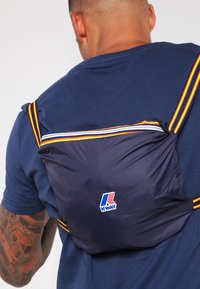 Námořnický nylonový batoh s oranžovými a bílými pruhovanými popruhy. Má logo K-WAY. Design je kompaktní a funkční, nošený na zádech.