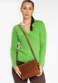 Borsa a tracolla in pelle marrone con finitura testurizzata e chiusura a patta, tenuta da una persona che indossa un cardigan verde a coste.