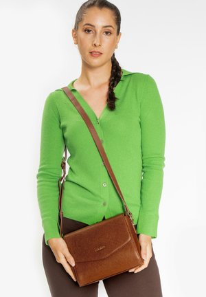Braune Leder-Umhängetasche mit strukturiertem Finish und Klappenverschluss, gehalten von einer Person in einer grünen Rippstrick-Cardigan.