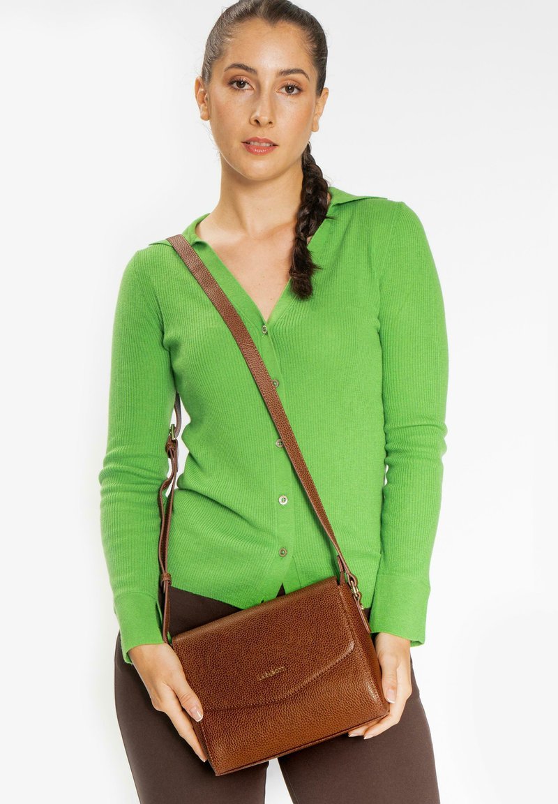 Borsa a tracolla in pelle marrone con finitura testurizzata e chiusura a patta, tenuta da una persona che indossa un cardigan verde a coste.