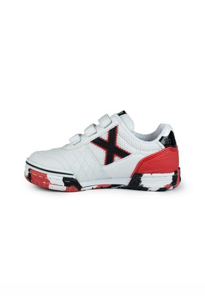 Zapatilla deportiva blanca con paneles de malla, acentos negros, contrafuerte rojo en el talón y suela pintada de negro y rojo. Cuenta con tres correas de gancho y lazo.