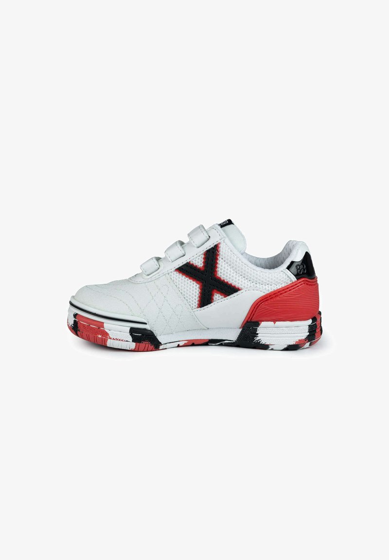 Zapatilla deportiva blanca con paneles de malla, acentos negros, contrafuerte rojo en el talón y suela pintada de negro y rojo. Cuenta con tres correas de gancho y lazo.