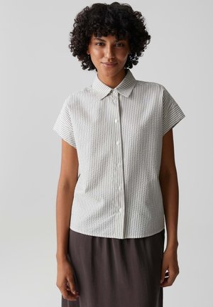 Femme aux cheveux bouclés portant une chemise blanche à manches courtes boutonnée avec de fines rayures verticales et une jupe foncée, debout devant un fond uni.