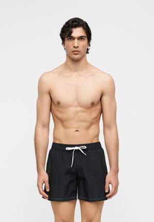 Póló nélküli férfi rövid göndör hajjal, fekete fürdőnadrágot visel fehér zsinórral, egyszerű világos háttér előtt áll.