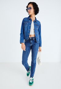 Niebieska kurtka dżinsowa na szarym crop topie, połączona z niebieskimi dżinsami i zielonymi sneakersami. Akcesoria to duże okulary przeciwsłoneczne i biała torebka.