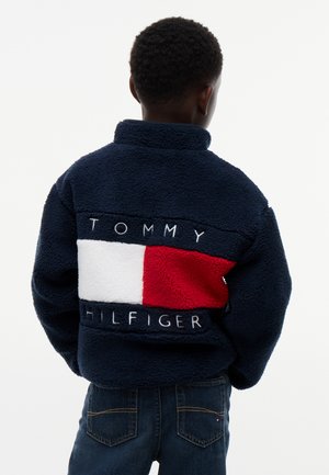 Παιδί που φοράει μπλε σκούρο fleece μπουφάν Tommy Hilfiger με μεγάλο κόκκινο, λευκό και μπλε σκούρο λογότυπο στην πλάτη, εμφανίζεται από πίσω.
