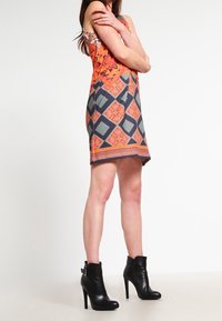 Robe sans manches colorée avec un motif géométrique orange et gris, associée à des bottines noires à talons hauts avec un détail à boucle.