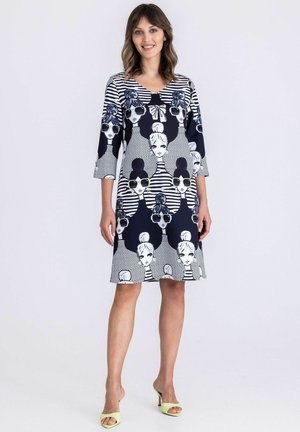 Frau trägt ein knielanges Kleid mit 3/4-Ärmeln, das einen schwarz-weißen grafischen Druck von Frauen mit Sonnenbrillen und gestreiften Mustern zeigt.