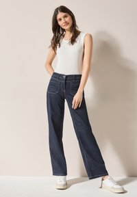 Jean en denim foncé à jambes larges avec des coutures contrastées, associé à un débardeur blanc ajusté. Le mannequin se tient sur un fond neutre.