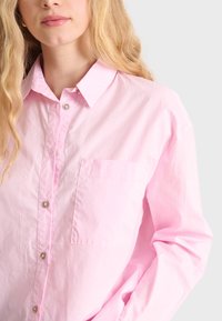 Light pink button-up shirt made of cotton, featuring a classic collar, front pocket, and shiny buttons. Long sleeves with soft texture.