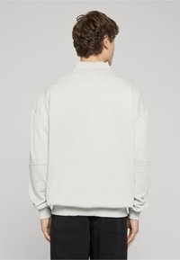 Muž s krátkými hnědými vlasy, oblečený v jednoduché bílé mikině a černých kalhotách, stojící zády k neutrálnímu šedému pozadí.