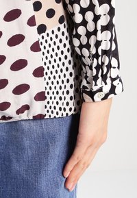 Blusa multi-pattern con pois neri, bianchi e bordeaux. Presenta un polsino con bottoni e una texture di tessuto morbida. Abbinata a un denim.
