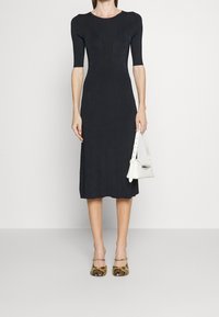 Robe noire ajustée à manches courtes, longueur genou, texture lisse. Associée à un sac à main blanc et des talons à imprimé serpent à motifs.