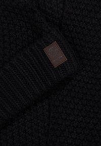 Czarny sweter z dzianiny z wyraźnym wzorem, ściągaczami przy mankietach oraz brązową, skórzaną metką z logo marki. Zbliżenie ukazuje szczegóły materiału.