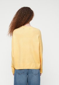 Femme aux cheveux longs et bouclés portant un pull jaune et un jean bleu, vue de dos sur un fond blanc uni.