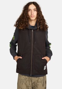 Schwarweste mit einem Reißverschluss vorne, zwei großen Taschen und einem Patch; getragen über einem grauen Hoodie mit grünen Blumenmustern an den Ärmeln.