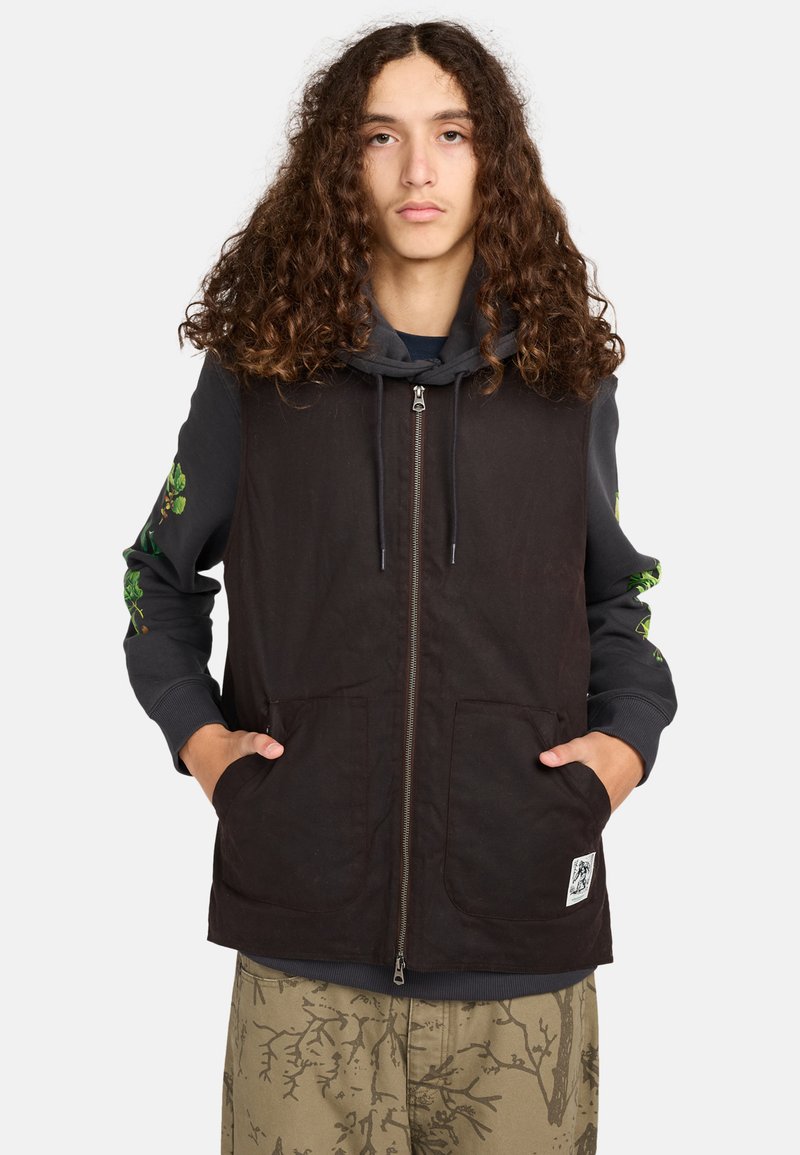 Schwarweste mit einem Reißverschluss vorne, zwei großen Taschen und einem Patch; getragen über einem grauen Hoodie mit grünen Blumenmustern an den Ärmeln.