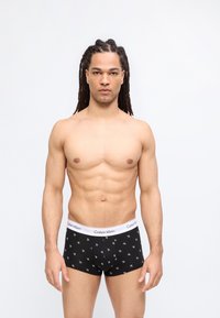 Γυμνασμένος άντρας με μακριά πλεξούδα στέκεται σε απλό φόντο, φορώντας μαύρα μποξεράκια Calvin Klein με λευκή ζώνη μέσης με λογότυπο.