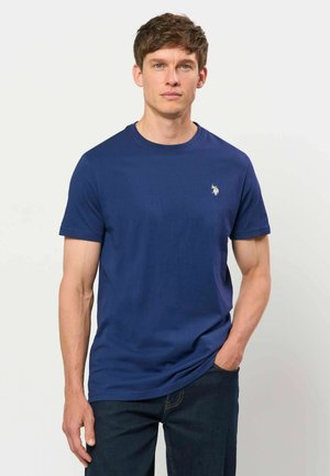 Mann trägt ein dunkelblaues T-Shirt mit kurzen Ärmeln und kleinem weißem besticktem Logo auf der linken Brust sowie dunkle Jeans, steht mit einer Hand in der Tasche.