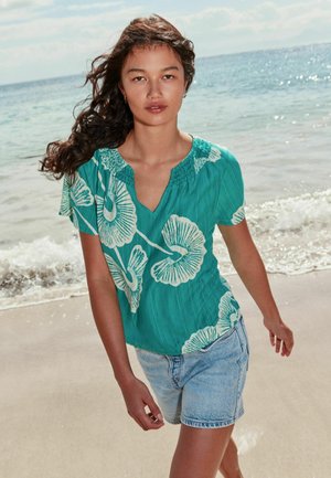 Frau mit langen dunklen Haaren, die ein türkisfarbenes Blumenoberteil und Jeansshorts trägt, geht an einem sonnigen Strand mit Ozeanwellen im Hintergrund entlang.
