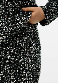 Robe noire recouverte de paillettes argentées brillantes, avec une coupe ajustée, une poche et des manches longues. La texture est réfléchissante et scintillante.