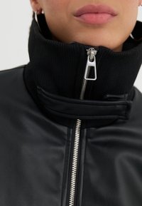 Chaqueta de cuero negra con un alto collar acanalado, detalle de cremallera plateada y líneas limpias. Textura suave con diseño ajustado.