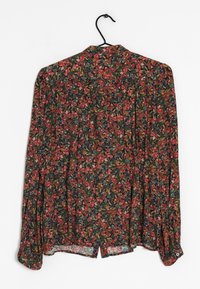 Chemisier à manches longues avec un motif floral rouge, vert et bleu, suspendu sur un cintre noir contre un fond blanc.