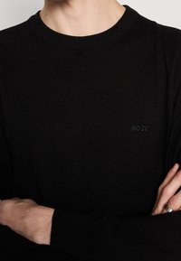 Personne portant une chemise noire à manches longues, bras croisés, montrant un petit logo "BOSS" sur la poitrine et une bague à la main droite.