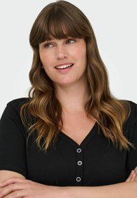 Wavy barna hajjal és frufruval rendelkező nő, fekete, gombos V nyakú pólót visel, oldalra néz, karba tett kézzel.